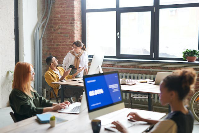 2 people are on computers in the foreground with 2 people in the background having a conversation