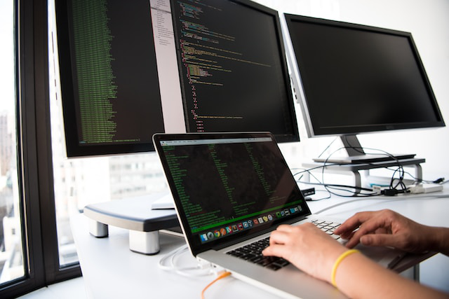 A person programming on a laptop with 2 monitors behind them